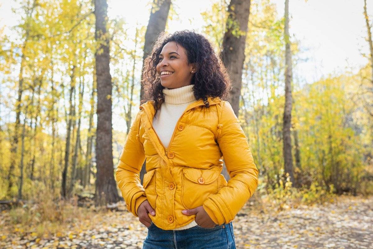 Smiling woman in a yellow puffer jacket and cream turtleneck standing in a sunlit autumn forest